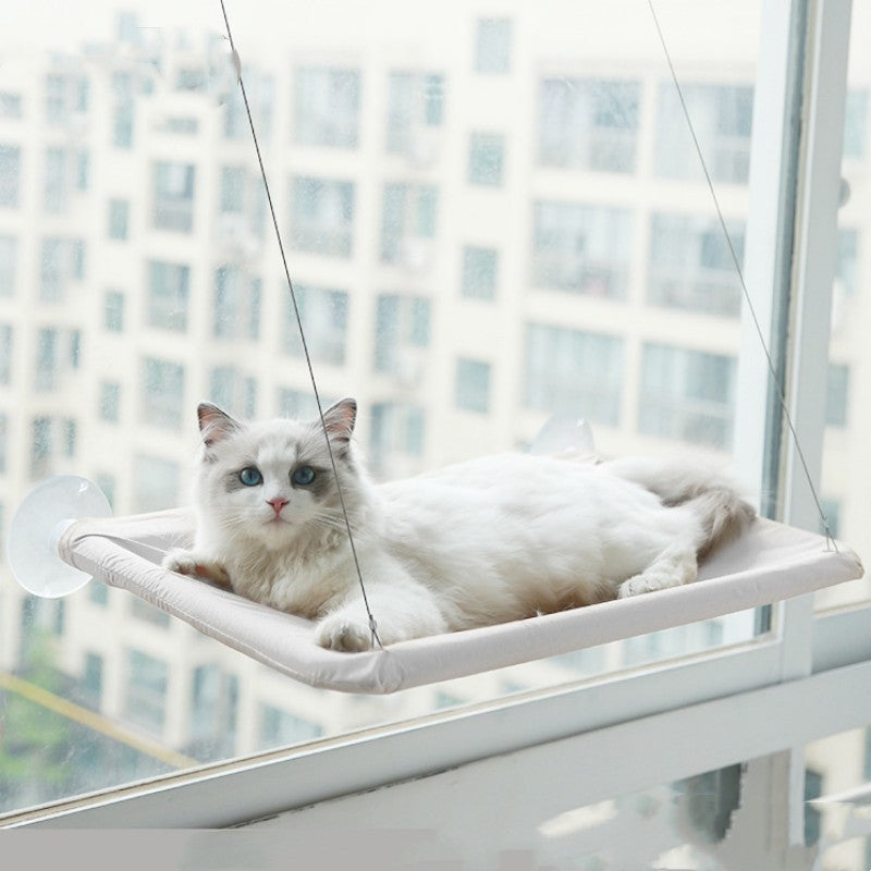 White cat resting on a hanging cat window hammock attached to a glass window with suction cups for birdwatching and sunbathing.