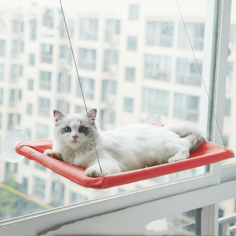 White cat resting on a red hanging cat window hammock attached with suction cups and ropes to a large window.