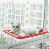 White cat resting on a red hanging cat window hammock attached with suction cups and ropes to a large window.